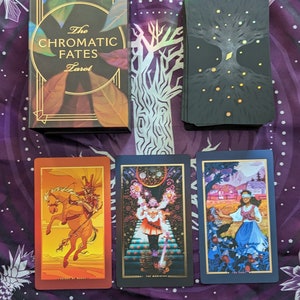 Może przedstawiać: Pudełko talii kart tarota z złotymi akcentami i tekstem "The Chromatic Fates Tarot". Trzy karty tarota ułożone są na fioletowym materiale z białym wzorem kwiatowym. Karty to "Rycerz Buław", "Mag" i "Głupiec".