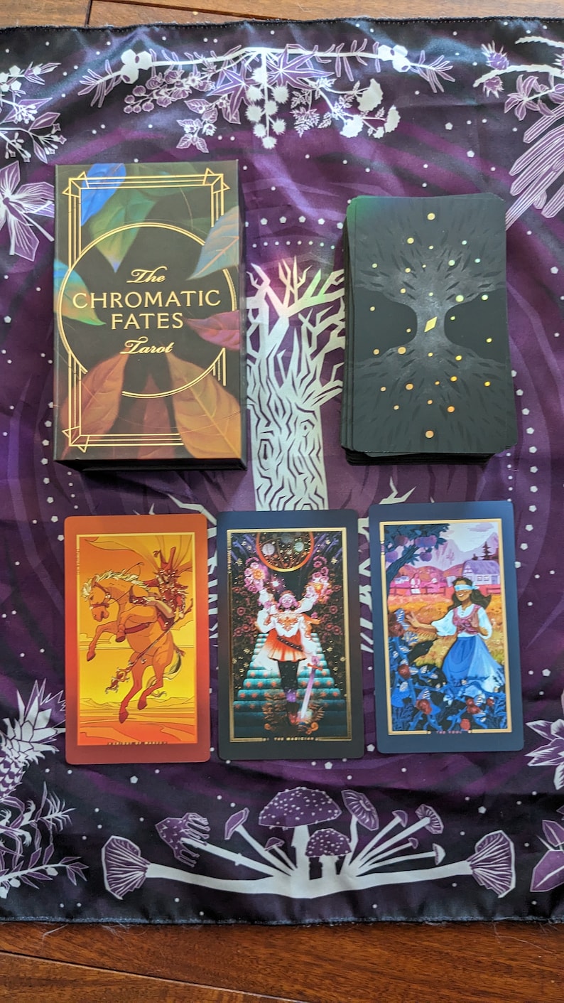 Może przedstawiać: Pudełko talii kart tarota z złotymi akcentami i tekstem "The Chromatic Fates Tarot". Trzy karty tarota ułożone są na fioletowym materiale z białym wzorem kwiatowym. Karty to "Rycerz Buław", "Mag" i "Głupiec".
