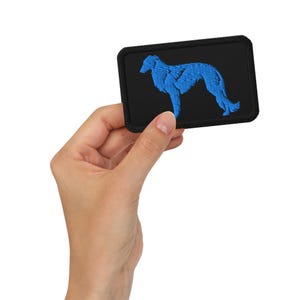 May include: A rectangular black patch with a blue embroidered image of a dog. The dog is a slender breed, possibly a Borzoi or similar, in a side profile. The patch has a black border.