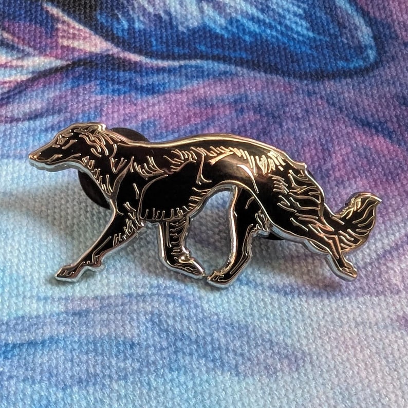 K&ouml;nnte beinhalten: Ein silberfarbener Ansteckpin mit einem stilisierten schwarzen Borzoi-Hund in Laufpose. Der Pin hat eine detaillierte Kontur und eine gl&auml;nzende schwarze Emaille-F&uuml;llung. Das Fell des Hundes ist strukturiert, vor blau-violettem Hintergrund.
