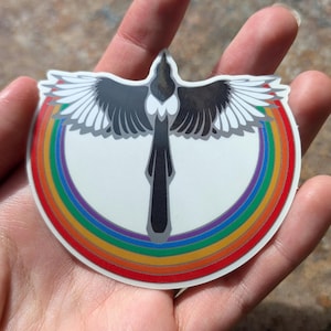 Puede incluir: Una urraca negra y blanca con las alas extendidas, volando sobre un arcoíris. El arcoíris es un círculo con los colores rojo, naranja, amarillo, verde, azul, añil y violeta.