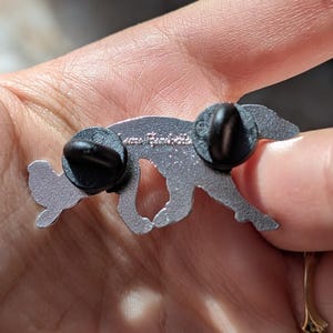 K&ouml;nnte beinhalten: Eine silberfarbene Metallnadel in Form eines laufenden Fuchses. Die Nadel hat zwei schwarze Nadelr&uuml;cken. Der Text "Anna Bondett" ist auf der R&uuml;ckseite der Nadel eingraviert. Der Fuchs hat einen kleinen Fl&uuml;gel auf dem R&uuml;cken.