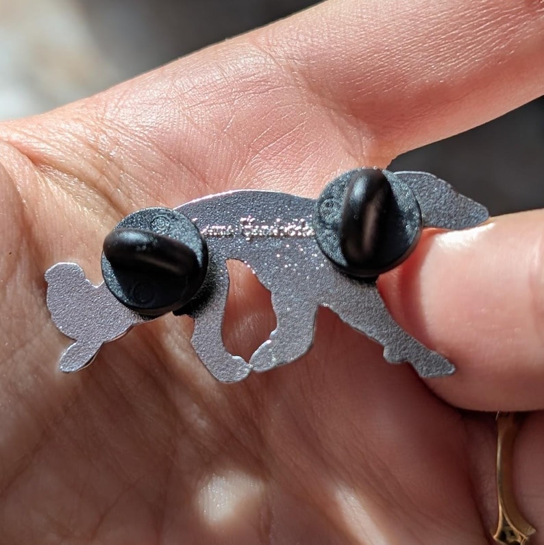 K&ouml;nnte beinhalten: Eine silberfarbene Metallnadel in Form eines laufenden Fuchses. Die Nadel hat zwei schwarze Nadelr&uuml;cken. Der Text "Anna Bondett" ist auf der R&uuml;ckseite der Nadel eingraviert. Der Fuchs hat einen kleinen Fl&uuml;gel auf dem R&uuml;cken.
