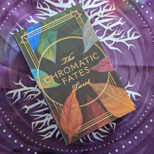 Może przedstawiać: Czarne pudełko na talię kart tarota z złotymi akcentami i tekstem "The Chromatic Fates Tarot" na przodzie. Pudełko jest ozdobione kolorowymi liśćmi.