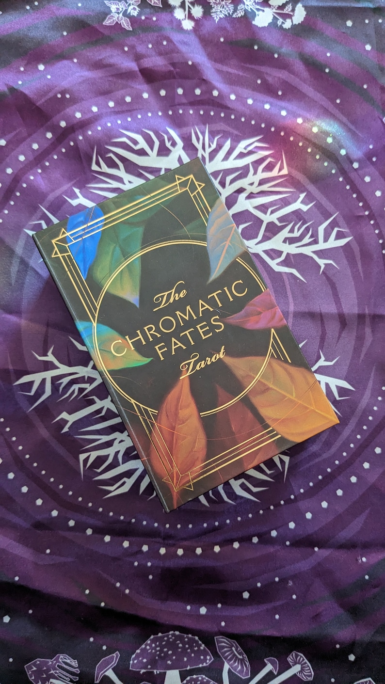 Może przedstawiać: Czarne pudełko na talię kart tarota z złotymi akcentami i tekstem "The Chromatic Fates Tarot" na przodzie. Pudełko jest ozdobione kolorowymi liśćmi.