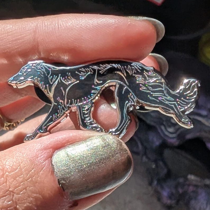 K&ouml;nnte beinhalten: Eine silberfarbene Anstecknadel in Form eines Hundes mit schwarzen Emaille-Details. Der Hund ist in einer Laufpose dargestellt, mit eingekr&auml;useltem Schwanz. Die Nadel hat ein gl&auml;nzendes, schillerndes Finish und wird von einer Person gehalten.