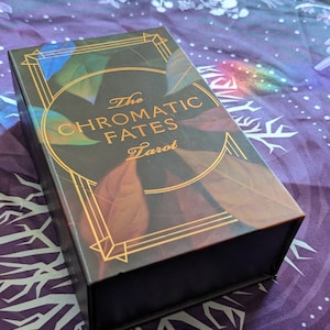 Może przedstawiać: Czarne pudełko z pozłacanymi napisami "The Chromatic Fates Tarot". Pudełko ma wz&oacute;r kolorowych liści z złotymi akcentami.