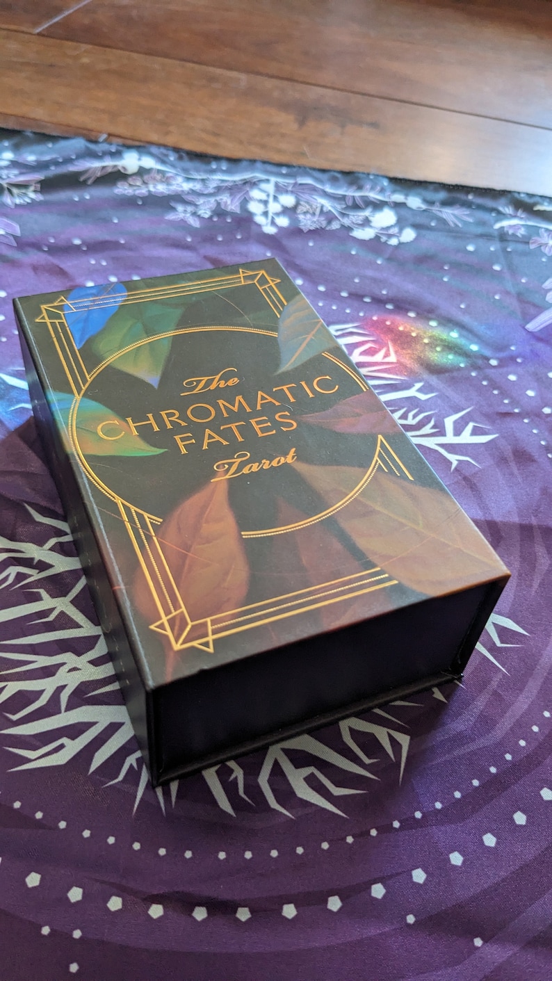 Może przedstawiać: Czarne pudełko z pozłacanymi napisami "The Chromatic Fates Tarot". Pudełko ma wz&oacute;r kolorowych liści z złotymi akcentami.