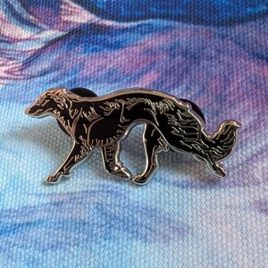 K&ouml;nnte beinhalten: Ein schwarz-silberner Emaille-Pin mit einem stilisierten Borzoi-Hund in einer Laufpose. Der Pin hat eine gl&auml;nzende silberne Umrandung und ein schwarzes Innenteil, vor einem blau-violetten Hintergrund.