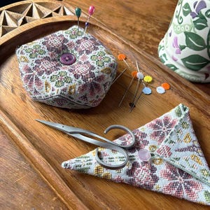 Puede incluir: Una bandeja de madera con un cojín de alfileres hexagonal, una pequeña bolsa de tela y unas tijeras de plata. El cojín de alfileres y la bolsa están decorados con motivos florales en tonos rosa, verde y blanco.