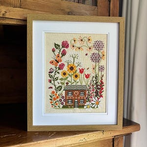 May include: A framed cross-stitch embroidery depicting a small cottage surrounded by a colourful garden with flowers, butterflies, and a heart-shaped window.