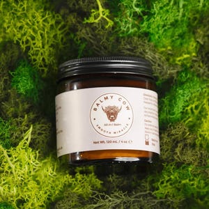 Puede incluir: Un tarro de vidrio marrón con tapa negra, etiquetado "Balmy Cow All-in-1 Balm Smooth Miracle". La etiqueta presenta una ilustración de un toro. El tarro contiene 120 ml de producto y está sobre musgo verde.