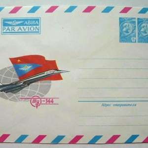 Pode incluir: Um envelope branco com listras azuis e rosa. O envelope tem uma bandeira vermelha, branca e azul com uma estrela. O envelope também tem uma imagem de um jato supersônico com o texto "Tu-144" e "Avia Par Avion".