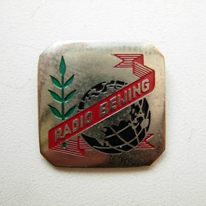 Peut inclure: Épinglette métallique argentée avec l'inscription "RADIO BEIJING" en rouge sur un globe noir et argenté. Une branche verte et une bannière rouge complètent le design. L'épinglette est de forme rectangulaire avec des coins arrondis.