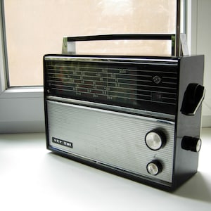 May include: A black and silver VEF 201 portable radio featuring a rectangular black casing, a silver mesh speaker grille, a top carrying handle, a telescopic antenna, a tuning dial with frequency markings, and two circular control knobs.