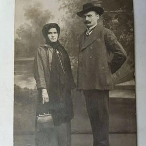 May include: Vintage black and white photograph of a man and woman. The man wears a double-breasted suit and a hat. The woman wears a long coat and holds a purse. The background features a blurred outdoor scene.