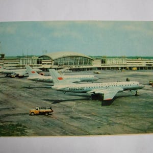 Op de afbeelding: Vintage ansichtkaart met verschillende passagiersvliegtuigen uit het Sovjettijdperk op een platform. De zilveren vliegtuigen hebben rode accenten en de luchthaventerminal is op de achtergrond te zien. Een gele vrachtwagen is ook aanwezig.