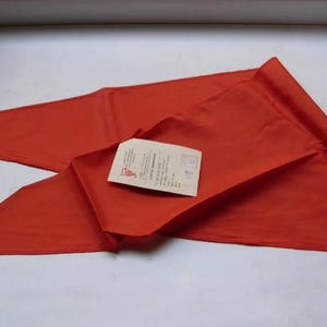 May include: A vibrant red triangular flag, folded and displayed on a white surface. A small rectangular tag with text is placed on top of the folded fabric. The flag has a pointed end and a folded edge.