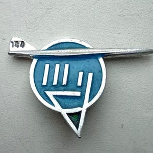 Pode incluir: Um alfinete vintage prateado com um design estilizado de avião. O alfinete tem um centro circular azul com linhas verticais brancas e um triângulo apontando para baixo. O número "144" é visível na asa do avião.