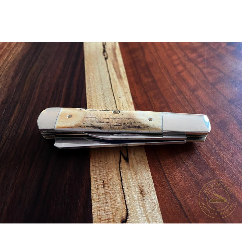 Puede incluir: Una navaja de bolsillo cerrada con mango de hueso y detalles met&aacute;licos plateados. La navaja est&aacute; sobre una superficie de madera con un patr&oacute;n de vetas marr&oacute;n oscuro y marr&oacute;n claro. La navaja tiene un dise&ntilde;o cl&aacute;sico y vintage.