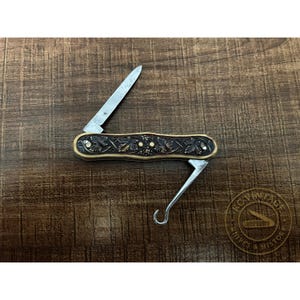 May include: A vintage pocket knife with a silver blade and a decorative handle. The handle features an ornate design with carved leaf and berry patterns. The knife also has a silver hook tool. The knife is displayed on a wooden surface.