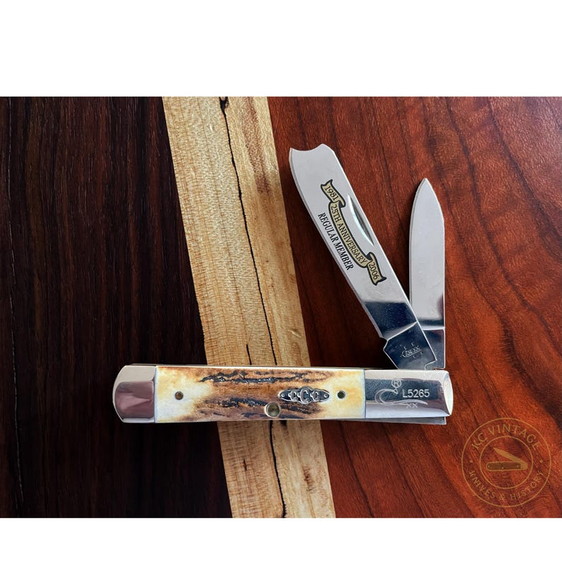 Puede incluir: Una navaja de bolsillo Case con mango de hueso y dos hojas abiertas. La navaja est&aacute; marcada con "25th Anniversary 2006 Regular Member" y "L5265". La navaja se muestra sobre una superficie de madera con vetas marr&oacute;n oscuro y marr&oacute;n claro.