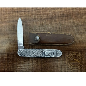 May include: A vintage pocket knife with a silver handle and a partially open blade. The handle features engraved names and a portrait. A brown leather sheath is next to the knife.