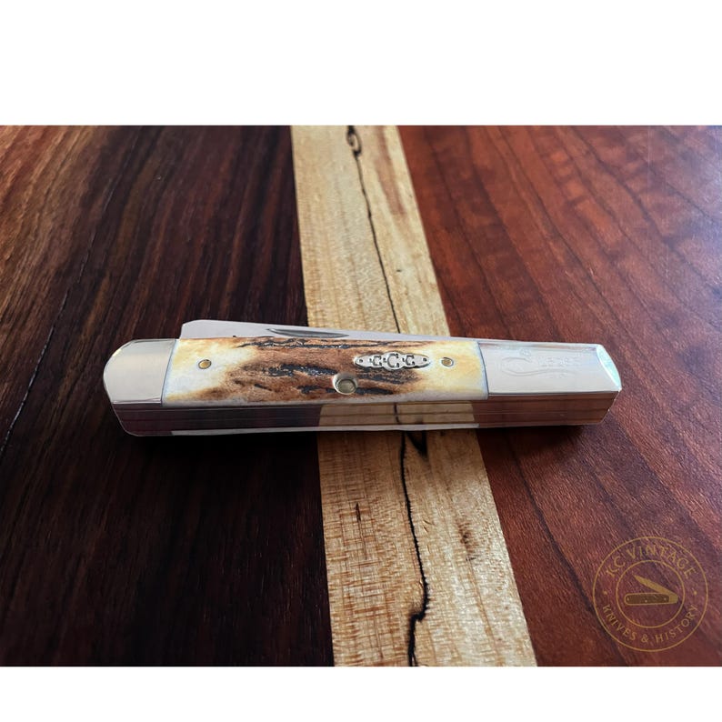 Puede incluir: Una navaja de bolsillo cerrada con mango de hueso y detalles met&aacute;licos plateados. La navaja tiene un logotipo de Case y las letras "G.C.G." son visibles en el mango. La navaja est&aacute; sobre una superficie de madera.