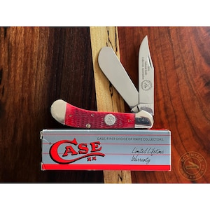May include: A red and silver Case pocket knife with two open blades, resting on a wooden surface. The knife has a red handle and a silver blade. The box below the knife reads "CASE. FIRST CHOICE OF KNIFE COLLECTORS."