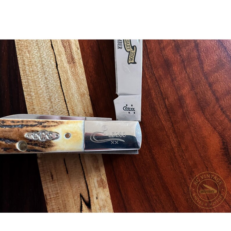 Puede incluir: Primer plano de una navaja de bolsillo con mango de color claro y hoja plateada. El mango tiene un dise&ntilde;o decorativo y la hoja est&aacute; marcada con "Case XX" y "LB265". La navaja est&aacute; sobre una superficie de madera.