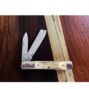 Puede incluir: Cuchillo de bolsillo vintage con dos hojas abiertas. El cuchillo tiene un mango de hueso con detalles plateados y una hoja de acero inoxidable. El cuchillo est&aacute; marcado "USA" y "5200PAZ". Se muestra sobre una superficie de madera.