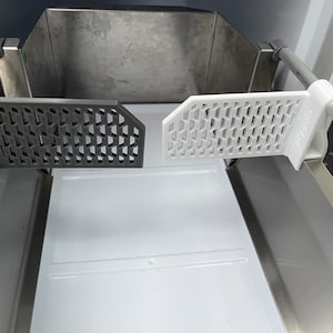 Puede incluir: Vista interior de una máquina de hielo con dos divisores de plástico perforados. Un divisor es de color gris oscuro, el otro es blanco. Los divisores están suspendidos sobre una bandeja de plástico blanco y componentes de acero inoxidable.