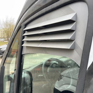 May include: A gray, angled window vent installed on a vehicle. The vent has multiple horizontal slats, providing a geometric design. The vehicle's interior, including the steering wheel, is visible through the window.