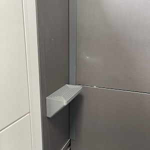 May include: A silver-coloured, fold-down step attached to a dark grey wall. The step has a ridged surface and is mounted on a vertical support. The wall is part of a larger structure, possibly a cabinet or a piece of furniture.