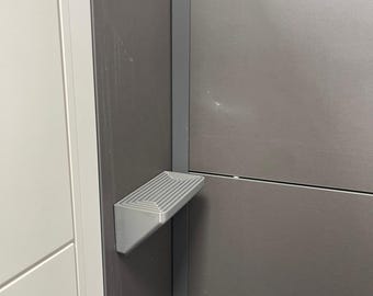 Op de afbeelding: Een zilverkleurige, opklapbare opstap bevestigd aan een donkergrijze muur. De opstap heeft een geribbeld oppervlak en is gemonteerd op een verticale steun. De muur maakt deel uit van een grotere structuur, mogelijk een kast of een meubelstuk.