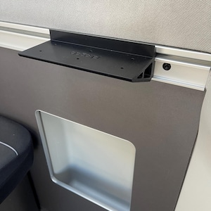 Puede incluir: Un estante de metal negro fijado a un riel plateado, montado en un panel gris oscuro. El estante tiene forma rectangular con un borde frontal ligeramente inclinado. El panel tiene un recorte rectangular con interior plateado.