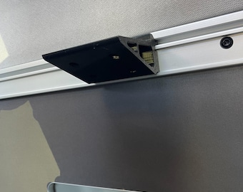May include: A black plastic bracket mounted to a silver track. The bracket is attached to a white wall.