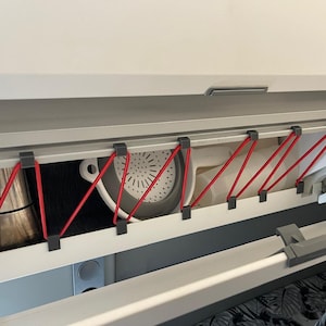 May include: A cabinet interior with various kitchen items secured by red bungee cords. Items include a metal coffee pot, a gray and white colander, and white utensils. The cabinet is white with gray accents.