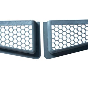 Op de afbeelding: Twee grijze ventilatieroosters met een honingraatpatroon. Elk rooster heeft een rechthoekig frame met een gebogen rand en een reeks zeshoekige openingen. De roosters zijn ontworpen om luchtstroom mogelijk te maken.