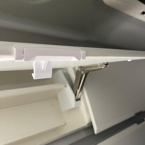 Può includere: Una clip di plastica bianca installata su una superficie bianca, probabilmente all'interno di un veicolo. La clip è rettangolare con un piccolo gancio. L'immagine mostra una vista parziale di un vano con una cerniera metallica.