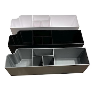 May include: Three desk organizers in white, black, and gray. Each organizer has multiple compartments of varying sizes, including a larger section on the left and smaller dividers on the right. The organizers are stacked on top of each other.