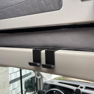 Op de afbeelding: Twee matzwarte J-vormige haken gemonteerd over de rand van een lichtbeige paneel, geplaatst direct onder een grijze matras met een zeshoekig gewatteerd patroon, in de cabine van een voertuig met een stuurwiel zichtbaar eronder.