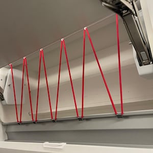 Op de afbeelding: Interieur van een witte kast met een rood bungee-koord systeem. De koorden zijn over de kast gespannen, bevestigd aan kleine witte clips. De kast heeft een zilveren scharnier en een wit interieur.