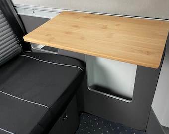 May include: A light brown bamboo table, approximately 60 cm long, attached to a dark grey interior wall. The table is rectangular and part of a camper van. A black seat with white trim is visible to the left, and a blue patterned floor is below.