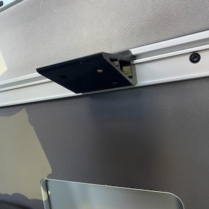 May include: A black plastic bracket mounted to a silver track. The bracket is attached to a white wall.