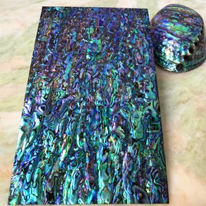 May include: A rectangular sheet of iridescent abalone shell inlay featuring a vibrant mosaic of deep blue, purple, and green hues, accompanied by a single rounded abalone shell with a matching multicolored, shimmering surface texture.