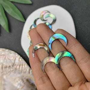 May include: Several crescent moon-shaped charms crafted from iridescent abalone shell. Each charm features a smooth, polished surface with a natural color gradient shifting between shades of green, blue, and purple, and includes a small hole.