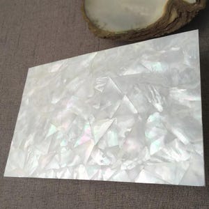 May include: A rectangular panel featuring a mosaic of white mother-of-pearl shell fragments. The surface is composed of numerous small, irregularly shaped, iridescent pieces fitted together to create a smooth, reflective, and shimmering finish.