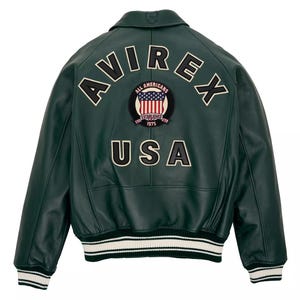 Puede incluir: Una chaqueta bomber de cuero verde oscuro con el texto AVIREX en letras grandes blancas y negras en la parte superior de la espalda. Debajo del texto hay un parche circular con un escudo y el texto ALL AMERICANS ESTABLISHED 1975. USA aparece debajo.