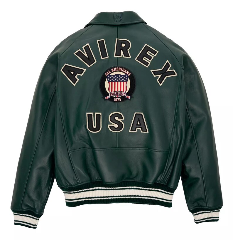Puede incluir: Una chaqueta bomber de cuero verde oscuro con el texto AVIREX en letras grandes blancas y negras en la parte superior de la espalda. Debajo del texto hay un parche circular con un escudo y el texto ALL AMERICANS ESTABLISHED 1975. USA aparece debajo.